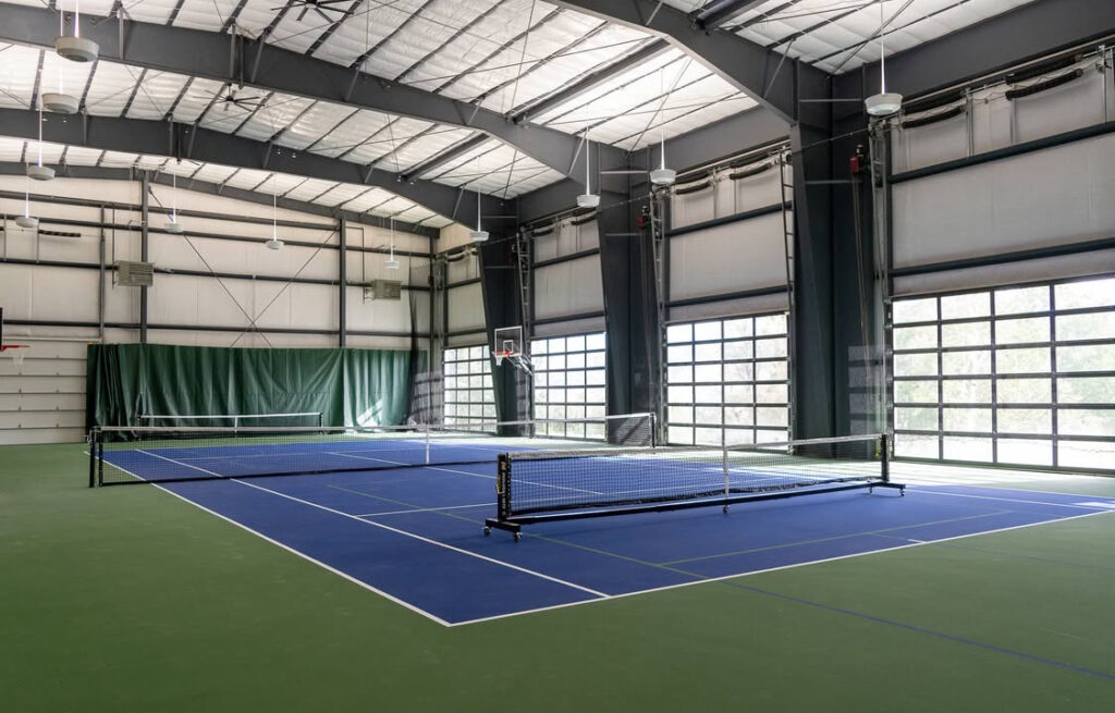 Indoor sports facility with high ceilings, featuring a blue tennis court and retractable basketball hoop, designed for versatile athletic activities.