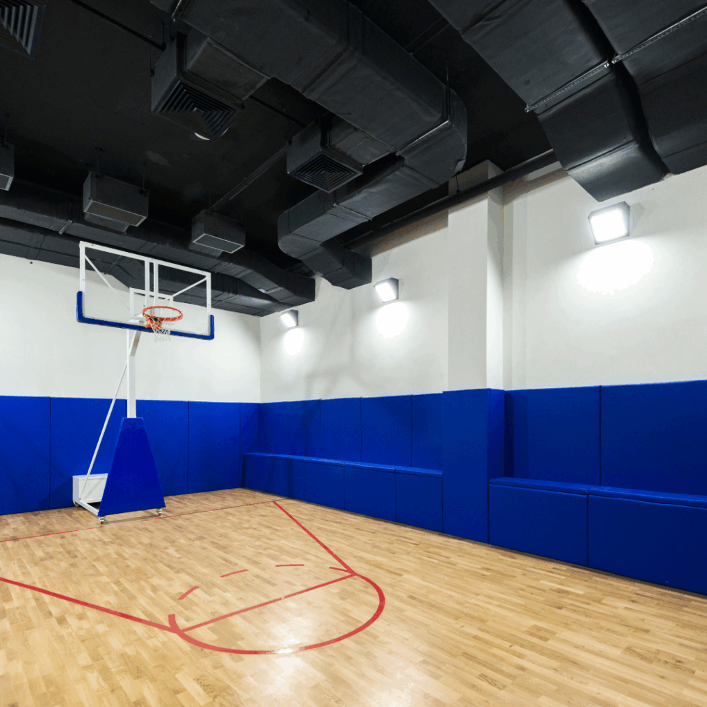 Indoor basketball court featuring hardwood flooring, blue wall padding for safety, and a professional hoop setup, emphasizing sports performance and aesthetics.