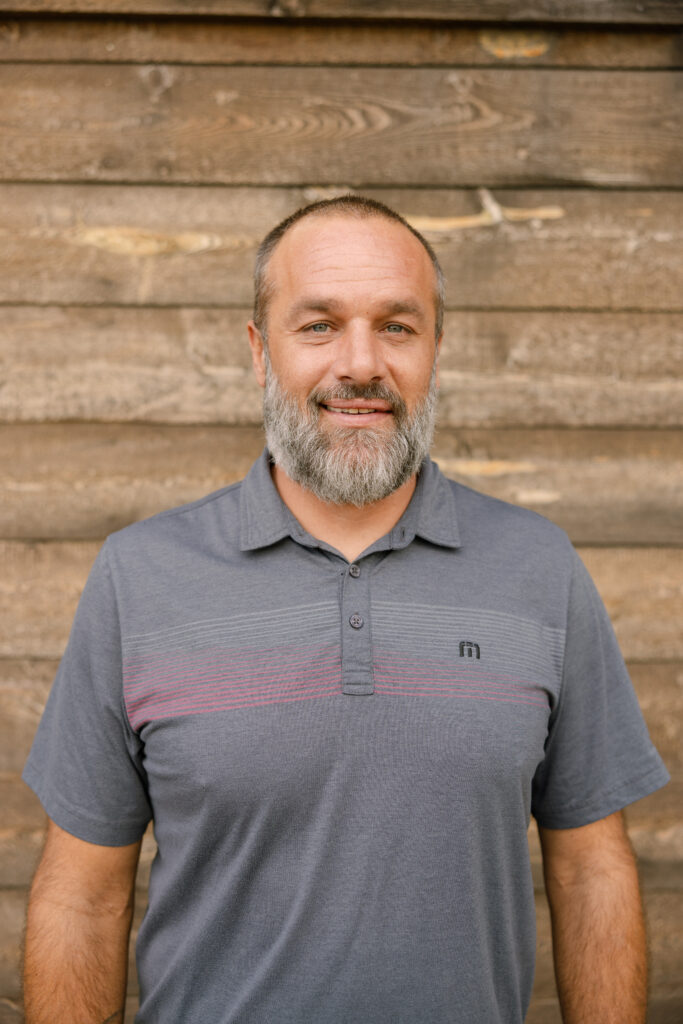 Camron Harper, craftsman with over 30 years of experience, smiling in front of wooden backdrop, showcasing dedication to craftsmanship and integrity at Peterson Builders.