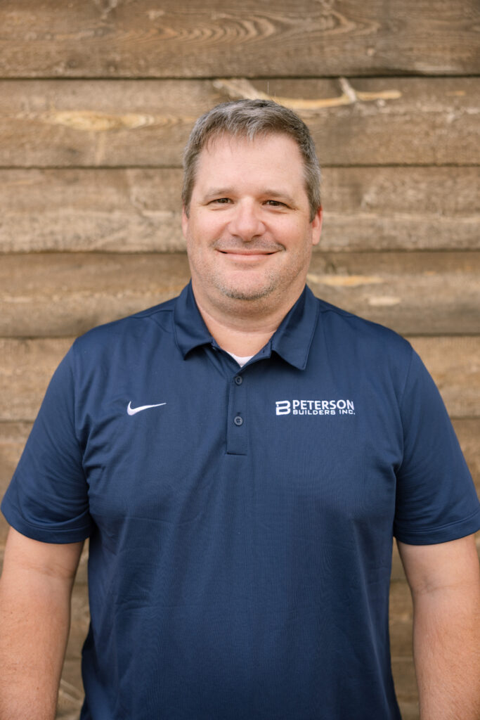 Jeremy Jones, Lead Estimator at Peterson Builders, smiling in a navy polo shirt with company logo against a wooden background.