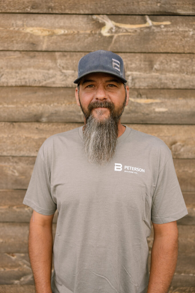 Robert Hultz, craftsman at Peterson Builders, wearing a gray t-shirt with logo, standing against a rustic wooden backdrop.