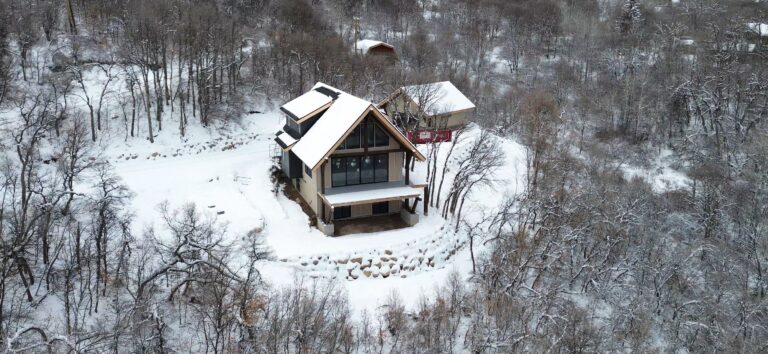 Aerial view of a modern A-frame home in a snowy landscape, surrounded by trees, showcasing winter construction progress for Peterson Builders' Huntsville project.